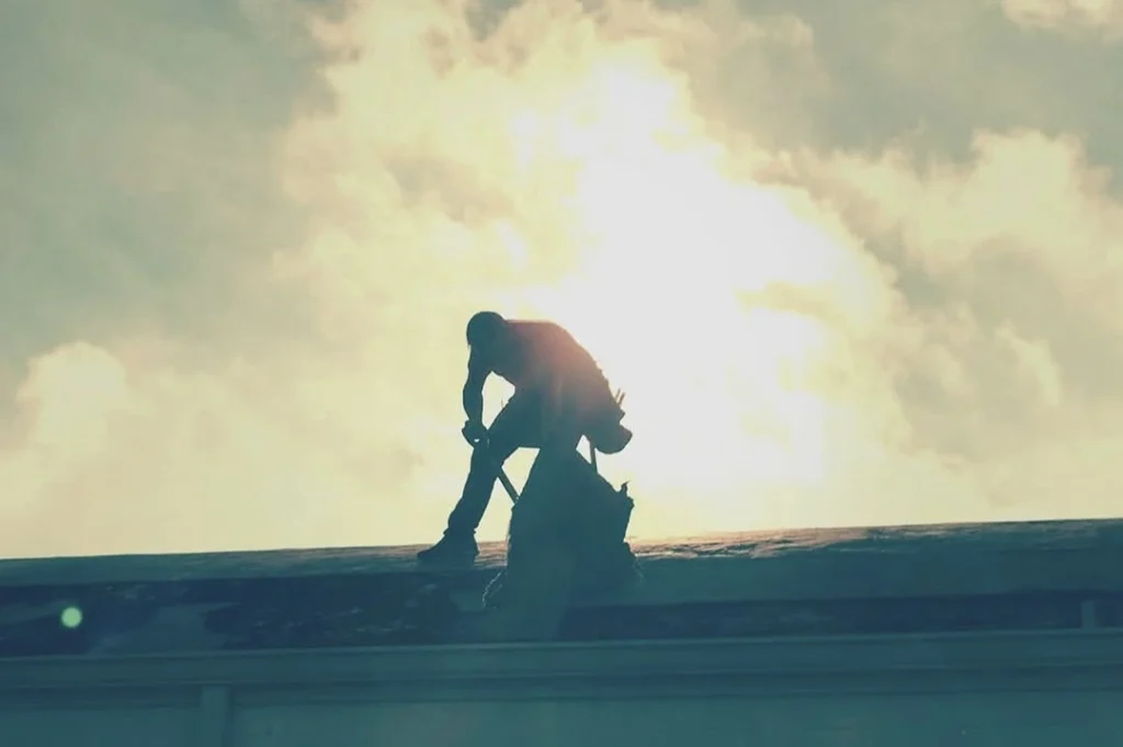 Silhouette of a roofing contractor working on a roof at sunrise with bright sunlight in the background.